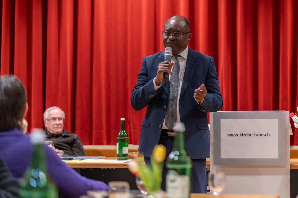 Ein Mann mit Glatze und blauem Anzug steht in einem Raum. In seiner rechten Hand hält er ein Mikrofon. 